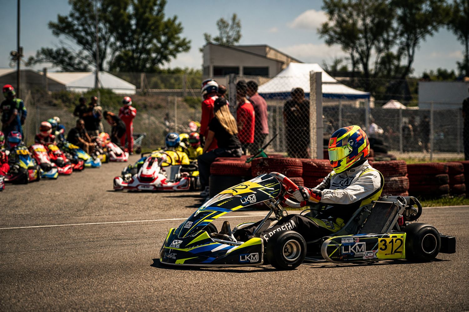 Kart LKM Racing n°312 en piste avec l'activité du paddock en arrière-plan