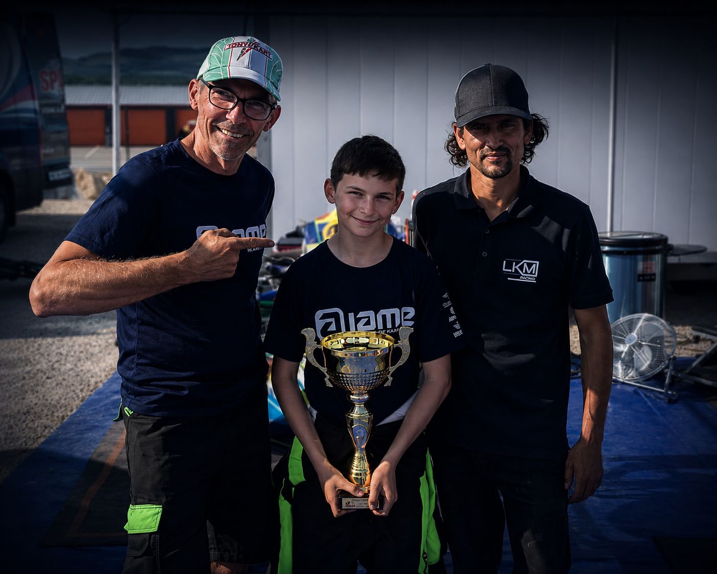 Un pilote LKM Racing récompensé d'un trophée, entouré de son coach et d'un membre de l'équipe