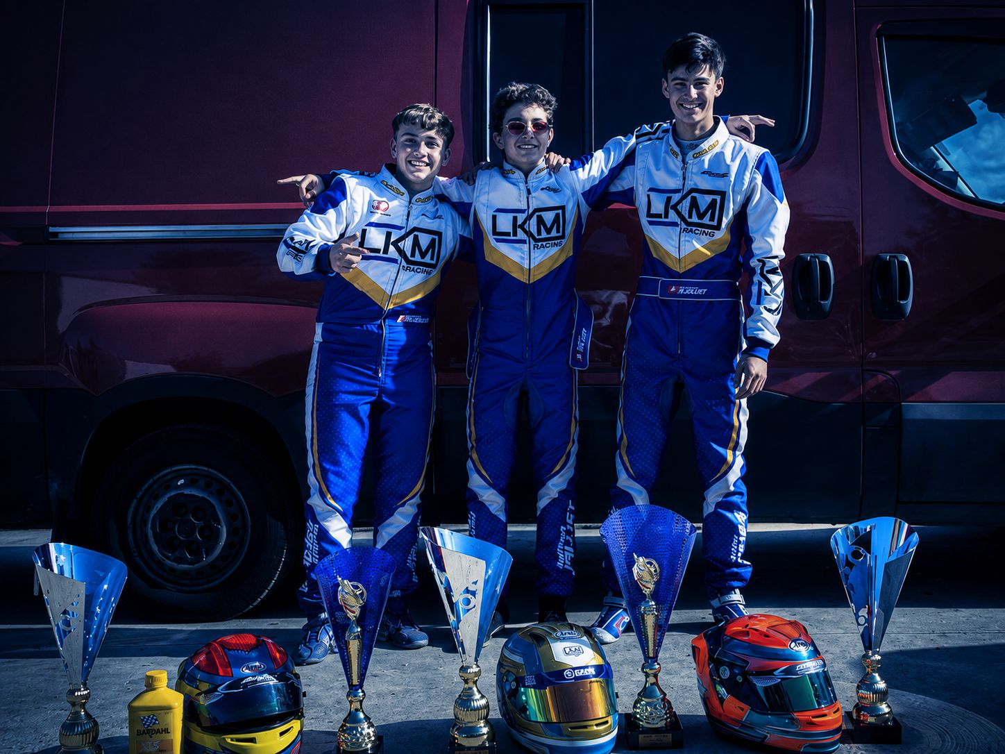 Trois pilotes LKM Racing devant le fourgon de l'équipe avec leurs trophées, casques et coupes